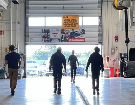 Cohort of 6 people in an automotive shop walking towards an open garage door